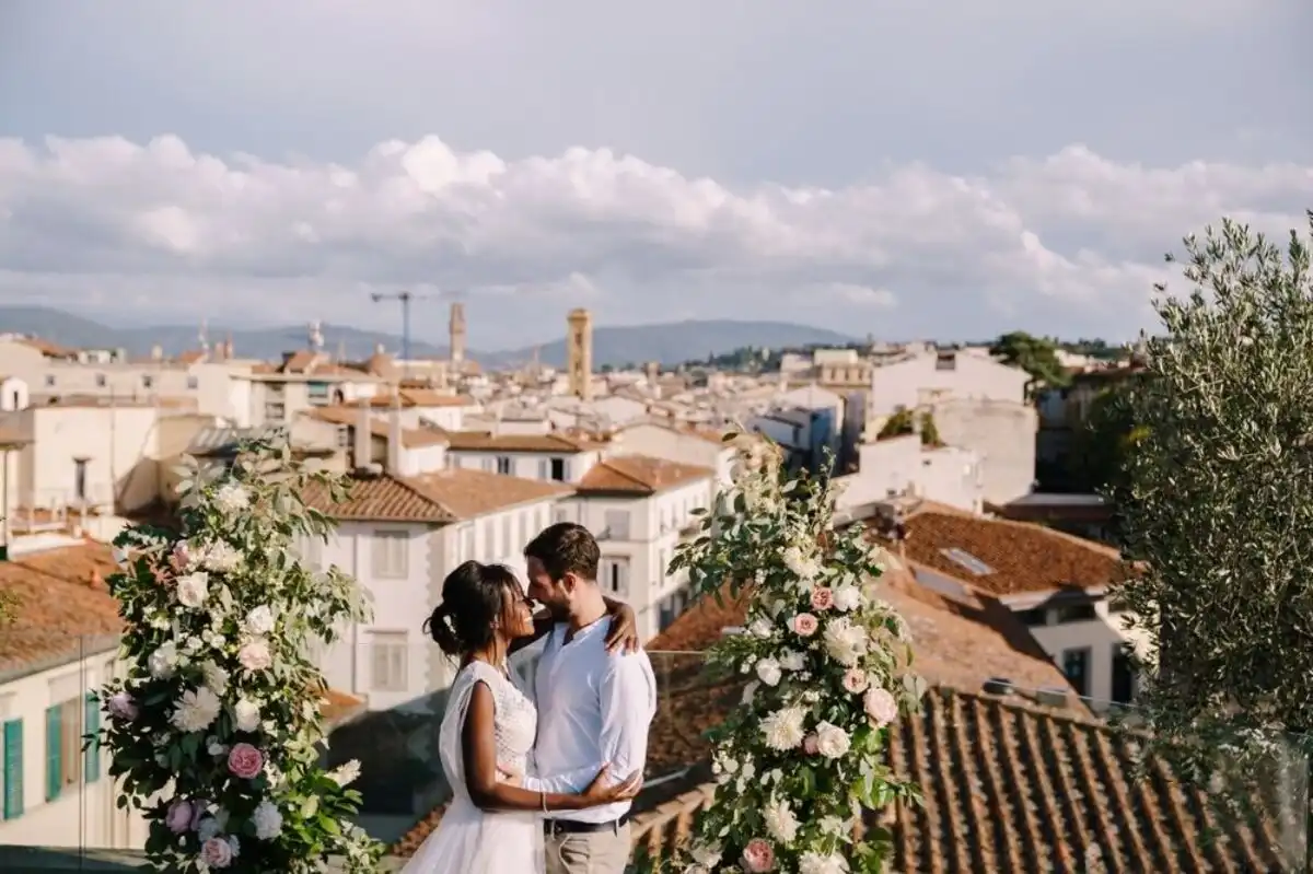 Casal apaixonado em um cenário romântico ao ar livre, cercado por flores e vista para uma cidade com telhados de cerâmica e céu nublado.