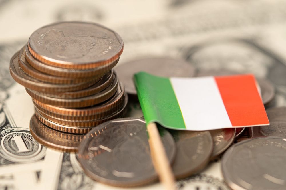Pilha de moedas com uma pequena bandeira da Itália ao lado, sobre uma cédula de dólar