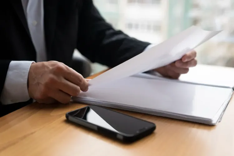 Pessoa em traje formal lendo documentos em papel em mesa de madeira com celular preto ao lado